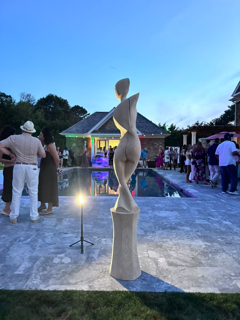 A carved wooden sculpture beside the pool as guests mingle at dusk during The Art Affair