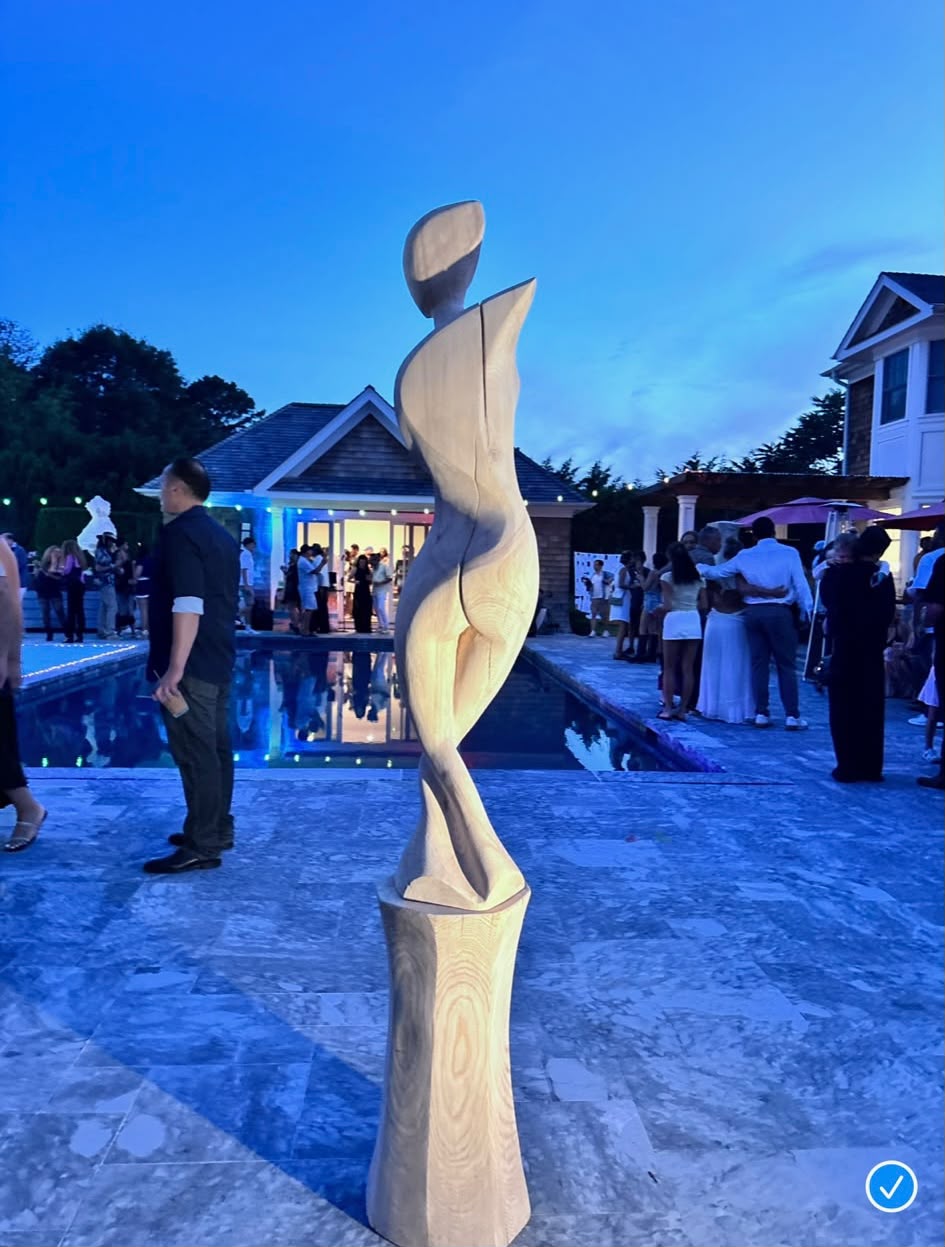 A carved wooden sculpture beside the pool as guests mingle at dusk during The Art Affair